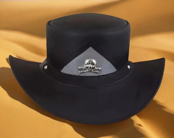 Puede incluir: Un sombrero negro con ala ancha y parte superior plana. Presenta un emblema plateado con un diseño de calavera y huesos cruzados. El sombrero está sobre un fondo arenoso.