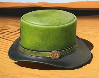 Puede incluir: Un sombrero de copa de cuero verde con ala negra y una banda decorativa. El sombrero presenta un botón dorado y está sobre un fondo desértico.