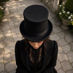 Puede incluir: Un sombrero de copa negro con ala ancha y cinta negra. El sombrero se lleva con un abrigo negro y una blusa de encaje. La imagen est&aacute; tomada desde arriba, mostrando el sombrero y la persona que lo lleva.