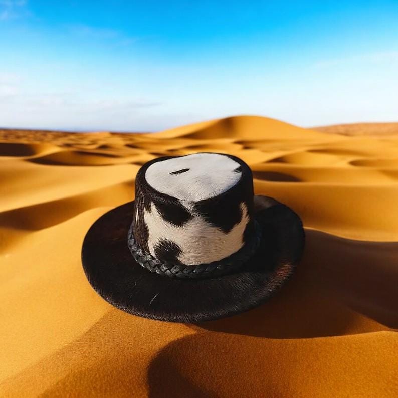 Op de afbeelding: Een zwart-witte koeienhuid hoed met een gevlochten band ligt op een zandduin. De hoed heeft een brede rand en een cilindrische kroon met een wit en zwart patroon. De achtergrond is een woestijnlandschap onder een blauwe lucht.