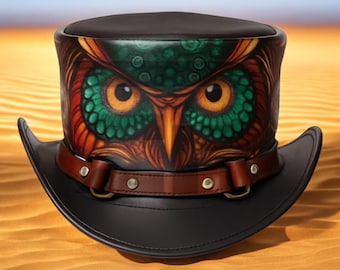 Puede incluir: Un sombrero de copa de cuero negro con una banda de cuero marrón y un diseño de búho. El búho tiene ojos naranjas, plumas verdes y naranjas, y está centrado en la corona del sombrero. El sombrero tiene un ala ancha.