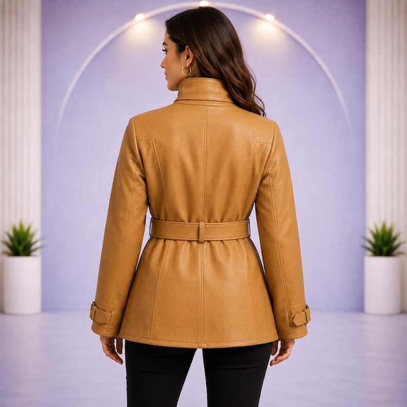 May include: A tan jacket with a high collar, long sleeves, and a matching belt at the waist. The jacket features vertical seam detailing on the back and adjustable straps with buckles on the cuffs, providing a structured fit and finish.