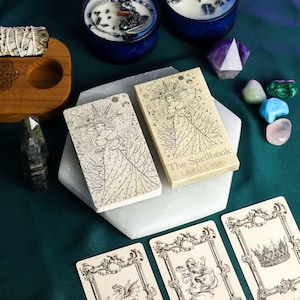 May include: A collection of spiritual items including oracle cards, crystals, candles, and sage. The oracle card box reads "The Spellbook Oracle Cards" with an illustration of a woman. The cards are on a white hexagonal surface.