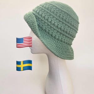 May include: A sage green crochet hat with a brim and textured stitch pattern. The hat is displayed on a white mannequin head. The hat's design includes a rounded crown and a slightly curved brim, offering a stylish and practical accessory.