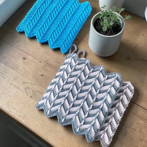 Pode incluir: Duas pegas de crochê feitas à mão. Uma é azul brilhante, a outra é cinza, branca e rosa claro. Ambas têm uma borda em zigue-zague e uma alça para pendurar. As pegas estão em uma superfície de madeira.
