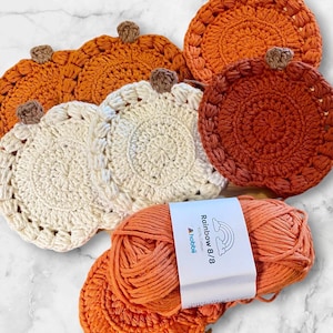 May include: A collection of round, pumpkin-shaped crochet coasters in shades of orange, cream, and rust, each featuring a small brown stem. A skein of orange Rainbow 8/8 100% cotton yarn by Hobbii with a white product label is positioned in the foreground.