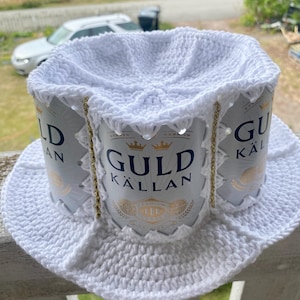 Puede incluir: Un sombrero blanco de ganchillo hecho a mano. La corona está formada por latas de cerveza Guld Källan recicladas, con detalles de cadena dorada. El ala es ancha y circular, un diseño original y reciclado.