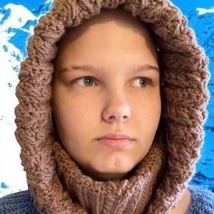 May include: A knitted hood and cowl set in a warm, earthy brown color. The hood features a textured, cable-knit design around the face, transitioning into a ribbed cowl. The background shows a blue sky and snow-covered mountains.