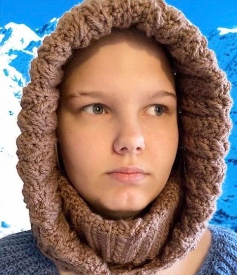 May include: A knitted hood and cowl set in a warm, earthy brown color. The hood features a textured, cable-knit design around the face, transitioning into a ribbed cowl. The background shows a blue sky and snow-covered mountains.