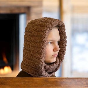 May include: A brown, knitted hood and scarf combination. The hood is pulled up, covering the head and neck. The knitted pattern is visible, and the color is a warm, earthy brown. The background includes a fireplace and wooden elements.