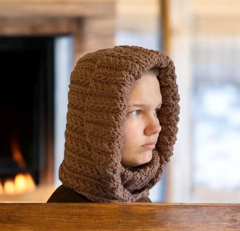 May include: A brown, knitted hood and scarf combination. The hood is pulled up, covering the head and neck. The knitted pattern is visible, and the color is a warm, earthy brown. The background includes a fireplace and wooden elements.