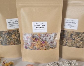 May include: Three brown paper pouches with clear windows, filled with dried herbs and flowers. The center pouch is labeled "Golden Glow Bath Soak." A bundle of sage, a crystal, and dried flowers are in front of the pouches. The products are for self-care.