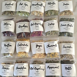 May include: A selection of small, clear plastic bags filled with dried herbs is shown from above. Each bag features a white label with the herb's name, like "Horsetail," "Hibiscus," and "Lavender," and the word "Organic." The herbs are diverse in colour and texture.