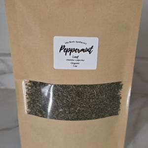 May include: A brown paper bag with a clear window, filled with dried peppermint leaves. The label reads "Peppermint Leaf" and "Organic 1 oz."