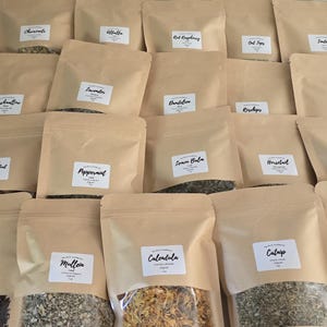 May include: A collection of twenty brown kraft paper pouches arranged in rows, each featuring a clear window at the bottom and a white rectangular label with black text identifying various dried herbs, flowers, and botanical ingredients.