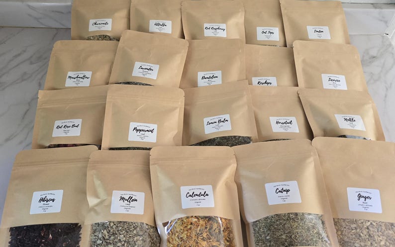May include: A collection of twenty brown kraft paper pouches arranged in rows, each featuring a clear window at the bottom and a white rectangular label with black text identifying various dried herbs, flowers, and botanical ingredients.