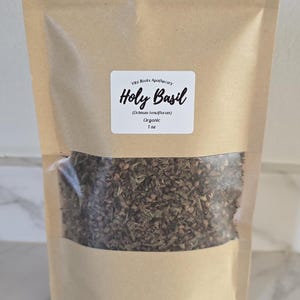 May include: A kraft paper bag with a clear window, filled with dried Holy Basil. A white label displays "Holy Basil" and "Organic 1 oz". The bag is sealed and sits on a marble surface.
