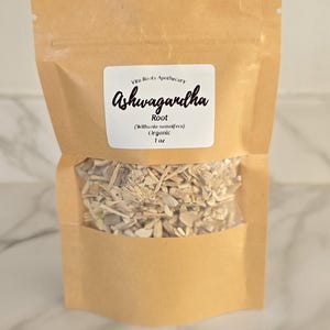 May include: A brown paper bag with a clear window, filled with dried root pieces. The bag has a white label with the text "Ashwagandha" in cursive, and the words "Root" and "Organic" are also visible.