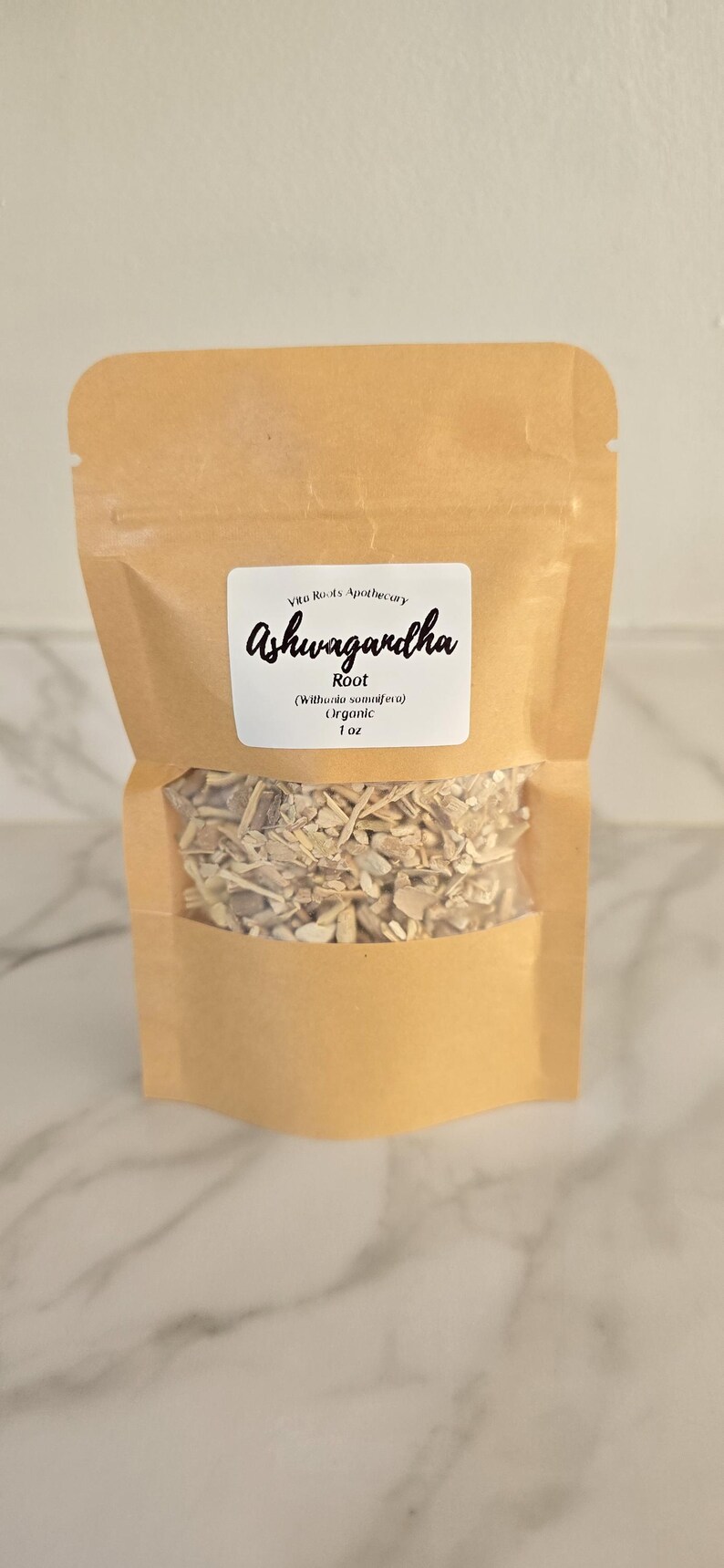 May include: A brown paper bag with a clear window, filled with dried root pieces. The bag has a white label with the text "Ashwagandha" in cursive, and the words "Root" and "Organic" are also visible.