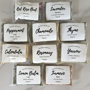May include: A selection of organic dried herbs in clear plastic bags. Each bag features a white label with the herb's name, including "Red Rose Bud," "Lavender," "Peppermint," and "Chamomile." The labels also display the botanical name and the word "Organic."