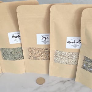 May include: Five light brown kraft paper stand-up pouches with clear windows, each containing dried herbs labeled Catnip, Horsetail, Ginger, Marshmallow Root, and Mullein Leaf. A small circular metal coin is placed in front of the pouches.
