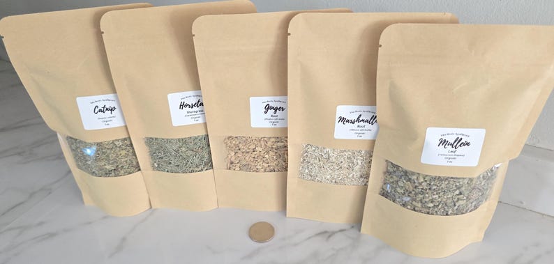 May include: Five light brown kraft paper stand-up pouches with clear windows, each containing dried herbs labeled Catnip, Horsetail, Ginger, Marshmallow Root, and Mullein Leaf. A small circular metal coin is placed in front of the pouches.