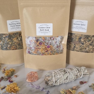 May include: Three brown paper pouches with clear windows displaying bath soaks and facial steam. The center pouch is labeled "Golden Glow Bath Soak" and contains dried flowers. A bundle of sage, a crystal, and dried flowers are in front.