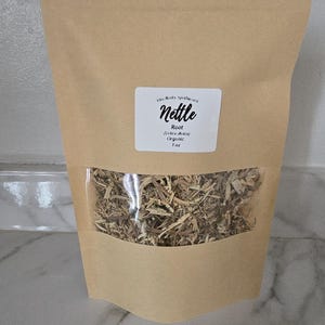 Könnte beinhalten: Ein brauner Papiersack mit einem transparenten Fenster, das getrocknete Brennnesselwurzel zeigt. Auf dem Etikett steht "Nettle Root (Urtica dioica) Organic 1 oz."