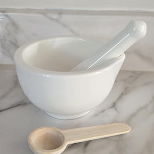 May include: A white ceramic mortar and pestle set with a smooth, glossy finish. The mortar is bowl-shaped with a wide opening, and the pestle is cylindrical with a rounded end. A small, light-colored wooden measuring spoon sits in front.
