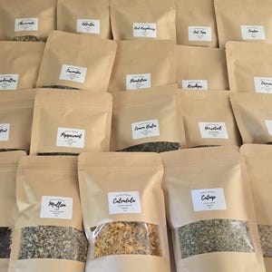 May include: A collection of twenty brown paper pouches containing dried herbs and botanicals. Each pouch features a clear rectangular window at the bottom and a white label with black text identifying the contents, including Hibiscus, Mullein, Calendula, Catnip, Ginger, Chamomile, Alfalfa, Red Raspberry, Oat Tops, Linden, Marshmallow, Lavender, Dandelion, Rosehips, Licorice, Red Rose Bud, Peppermint, Lemon Balm, Horsetail, and Nettle.