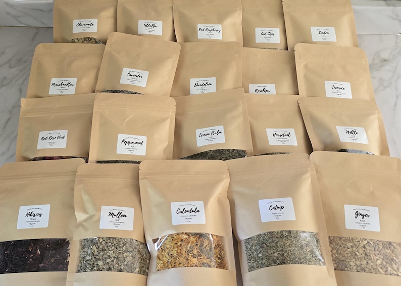 May include: A collection of twenty brown paper pouches containing dried herbs and botanicals. Each pouch features a clear rectangular window at the bottom and a white label with black text identifying the contents, including Hibiscus, Mullein, Calendula, Catnip, Ginger, Chamomile, Alfalfa, Red Raspberry, Oat Tops, Linden, Marshmallow, Lavender, Dandelion, Rosehips, Licorice, Red Rose Bud, Peppermint, Lemon Balm, Horsetail, and Nettle.
