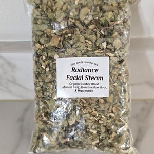 Puede incluir: Una bolsa de plástico transparente llena de hierbas verdes secas. Una etiqueta blanca en la bolsa dice "Vita Roots Apothecary Radiance Facial Steam" con los ingredientes: hoja de gordolobo, raíz de malvavisco y menta.