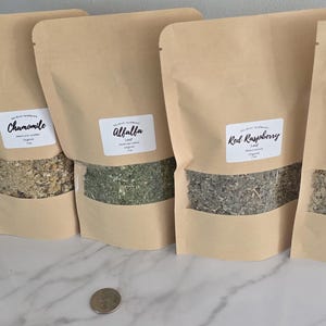 May include: Five stand-up pouches made of light brown kraft paper, each featuring a clear window displaying dried herbs. The white labels are printed with the text Peppermint, Chamomile, Alfalfa, Red Raspberry, and Linden. A US quarter is visible.