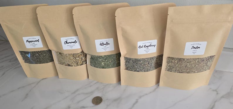 May include: Five stand-up pouches made of light brown kraft paper, each featuring a clear window displaying dried herbs. The white labels are printed with the text Peppermint, Chamomile, Alfalfa, Red Raspberry, and Linden. A US quarter is visible.