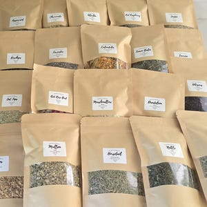 May include: A collection of twenty brown paper pouches containing various dried herbs and botanicals. Each pouch features a clear window at the bottom to display the contents and a white label with the name of the herb, including Peppermint, Chamomile, Alfalfa, Red Raspberry, Linden, Rosehips, Lavender, Calendula, Lemon Balm, Damiana, Oat Tops, Red Rose Bud, Marshmallow, Dandelion, Hibiscus, Ginger, Mullein, Horsetail, Nettle, and Catnip.