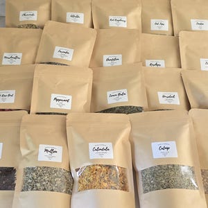 May include: A collection of twenty light brown paper pouches arranged in rows, each featuring a white rectangular label with black text identifying various dried herbs and botanicals, including hibiscus, calendula, and peppermint, with clear windows.