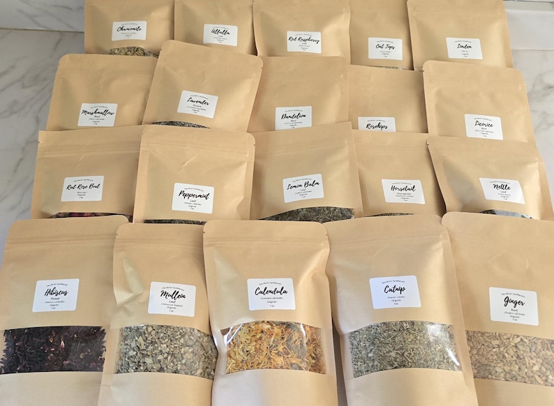 May include: A collection of twenty light brown paper pouches arranged in rows, each featuring a white rectangular label with black text identifying various dried herbs and botanicals, including hibiscus, calendula, and peppermint, with clear windows.