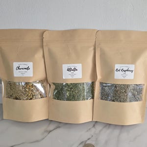 May include: Three light brown stand-up pouches containing dried herbs, each with a clear window and a white label. The labels read Chamomile, Alfalfa, and Red Raspberry. Each pouch is marked as 1 oz. A silver coin is placed in front of the pouches.