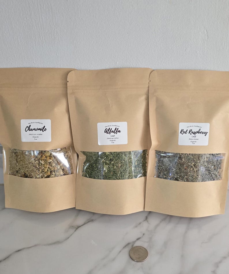May include: Three light brown stand-up pouches containing dried herbs, each with a clear window and a white label. The labels read Chamomile, Alfalfa, and Red Raspberry. Each pouch is marked as 1 oz. A silver coin is placed in front of the pouches.