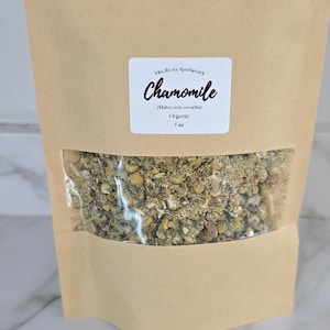 May include: A brown paper bag with a clear window displaying dried chamomile flowers. A white label on the bag reads "Chamomile" with the botanical name and "Organic" below. The bag contains 1 oz of chamomile.
