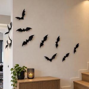 Pode incluir: Uma coleção de adesivos de parede em forma de morcego preto dispostos em um padrão de voo em uma parede de cor clara. O conjunto inclui várias silhuetas individuais de morcegos de tamanhos e posições de asas variadas.