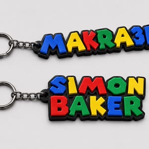 Pode incluir: Dois chaveiros com letras coloridas em relevo. O chaveiro superior diz "MAKRA3D" em letras azuis, vermelhas, verdes, amarelas e azuis com uma borda preta. O chaveiro inferior diz "SIMON BAKER" em letras azuis, amarelas, vermelhas, verdes e azuis com uma borda preta.