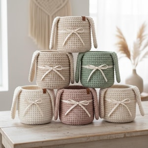 May include: A stack of six crochet baskets in various colors: cream, beige, sage green, and dusty rose. Each basket has bunny ear accents, a ribbon bow, and a small leather tag. The baskets are stacked on a wooden surface.