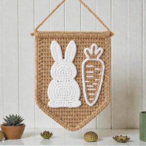 May include: A tan crocheted Easter banner featuring a white bunny and carrot design. The banner has a rectangular shape with a pointed base and a rope for hanging. The bunny and carrot are outlined in white.