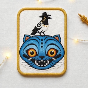 May include: Embroidered patch featuring a blue cat with orange eyes and sharp teeth, topped by a black and white bird wearing a hat. The patch has a white background with a gold border, showcasing a whimsical, illustrative design.