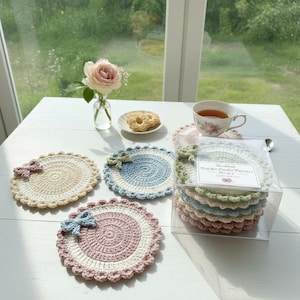 Op de afbeelding: Handgemaakte gehaakte onderzetters in pastelkleuren, waaronder crème, blauw en roze. Elke onderzetter heeft een spiraalvormig ontwerp met een decoratieve strik. Een set onderzetters wordt gepresenteerd in een transparante doos, samen met een kopje thee en een klein bord.