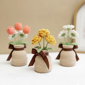 Pode incluir: Três arranjos de flores de crochê em vasos bege com laços de fita marrons. As flores incluem tulipas pêssego, rosas amarelas e margaridas brancas. Os vasos têm um padrão texturizado e as flores têm caules e folhas verdes.