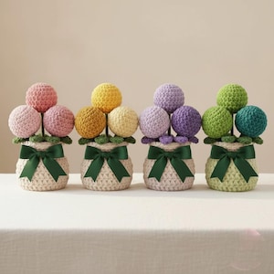 Puede incluir: Cuatro arreglos florales de ganchillo en macetas color crema. Cada maceta tiene un lazo verde y contiene flores esféricas en tonos rosa, amarillo, morado y verde. Las flores están dispuestas sobre tallos verdes.