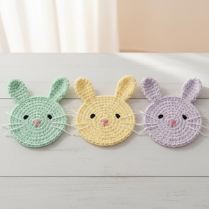 Może przedstawiać: Trzy ręcznie robione podkładki pod kubki w kształcie królika, w pastelowych kolorach: miętowym, żółtym i lawendowym. Każda podkładka ma okrągły kształt z uszami królika, czarnymi oczami, różowym nosem i białymi wąsami.