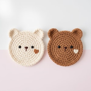 May include: Two crocheted bear-shaped coasters. One is cream-colored with a brown heart, the other is brown with a white heart. Both have black eyes and brown noses. The coasters are circular with small, rounded ears.